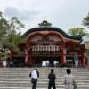 Shinto shrine with a red wooden façade, curved green roof, stone steps, and fox statues at the entrance; visitors ascend to the main hall.